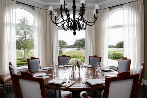 Interesting-dining-room-with-curving-windows-and-green-view-and-wooden-round-table-and-antique-chandelier-and-glass-cup-and-plate